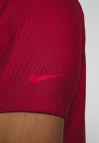 Röd strukturerad sportskjorta med kort ärm, prydd med en rosa Nike-logotyp på överarmen. Materialet verkar vara andningsbart och sportigt.