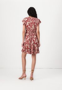 Femme aux longs cheveux bouclés portant une robe courte à manches courtes rouge et blanche à motifs floraux et des sandales beiges à talons, photographiée de dos sur un fond uni.