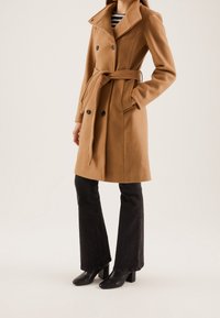 Kamelinvillaisen sekoitteen takki, jossa kaksirivinen etuosa, ylisuurikokoinen kaulus ja vyötäröllä vyö. Yhdistetty liehuviin mustiin farkkuihin ja mustiin korkokenkiin.