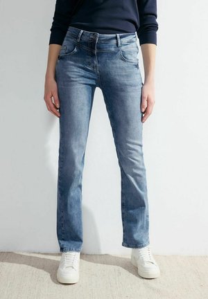 Persona que lleva jeans rectos de color azul claro, zapatillas blancas y una blusa de manga larga de color azul marino oscuro, de pie sobre una alfombra beige.