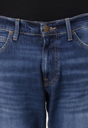 Close-up af blå denimjeans med messing Lee-knap, synlige bæltestropper, frontlommer og orange syninger på sort baggrund.