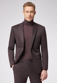 Brązowy dwuczęściowy garnitur z dopasowaną marynarką i pasującymi spodniami, noszony na ciemnym golfie. Zawiera kieszeń i subtelny detal z logo.