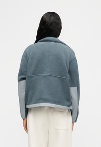 Άτομο με μακριά σκούρα μαλλιά φοράει μπλε-γκρι φλις μπουφάν με ανοιχτό γκρι πατς στους αγκώνες και κρεμ παντελόνι, ορατό από πίσω.