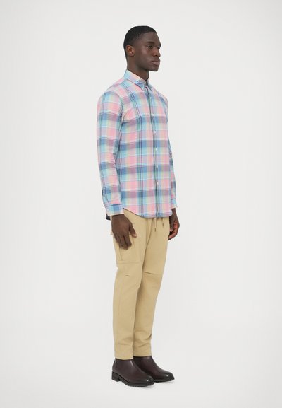 Ozka srajca iz kariranega blaga z dolgimi rokavi v rožnati, modri in beli barvi, z ovratnikom z gumbki. Kombinirana z bež hlačami in rjavimi škornji.