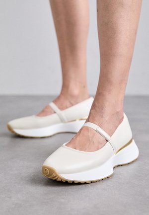 Stopy w beżowych butach w stylu Mary Jane z grubymi białymi podeszwami i złotym akcentem na pięcie, stojące na szarej podłodze.