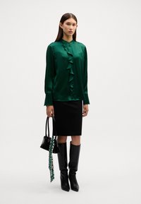 Dunkelgrüne Satinbluse mit Rüschendetails auf der Vorderseite, langen Ärmeln und einem schwarzen Bleistiftrock. Das Model hält eine schwarze Handtasche mit einem gemusterten Schal als Accessoire.