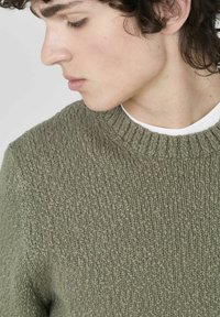 Pull en maille vert olive avec une finition texturée, col rond côtelé, et une chemise blanche superposée en dessous. La vue rapprochée met en avant le détail du tissu.
