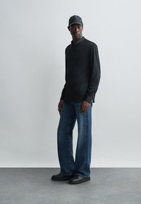 Musta pitkähihaine paita, vaalean siniset suorat farkut, mustat lenkkarit ja musta lippalakki, joita henkilö käyttää seisten harmaata taustaa vasten.