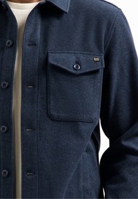 Marineblauwe shirtjack van wolmix met knoopsluiting, enkele borstzak en contrasterende zwarte knopen. Zachte textuur en getailleerde pasvorm.
