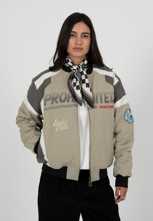 Kvinde iført beige og grå racerkørerjakke med teksten "FORBUDT RACING", ternet halstørklæde, sorte bukser og hvid skjorte, stående mod en hvid baggrund.