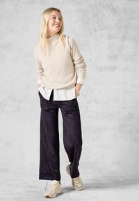 Beige geribde gebreide trui over een witte blouse met kraag, gecombineerd met wijde zwarte broeken en beige sneakers. Soepele texturen en een relaxte pasvorm.