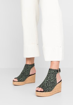 Sandales compensées en tissu imprimé léopard vert et noir, avec orteils ouverts et talons, portées avec un pantalon blanc retroussé.