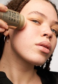 En gullfarget, sylindrisk stift merket "BLUR STICK BRIGHT" påføres kinnet. Subjekten har krøllete hår og ansiktspiercinger.