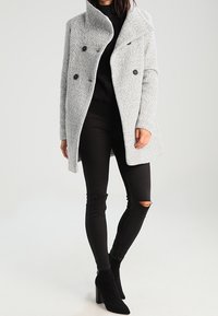 Grauer doppelt knöpfbarer Mantel mit strukturiertem Finish und großem Kragen, getragen über einem schwarzen Pullover und schwarzen zerrissenen Jeans, mit schwarzen Stiefeletten.
