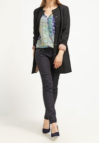 Musta pitkä takki kuvioidun sinivihreän paidan päällä, yhdistettynä tummiin istuviin housuihin ja laivastonsinisiin korkeakorkoisiin kenkiin. Sileä kangas ja räätälöity istuvuus.