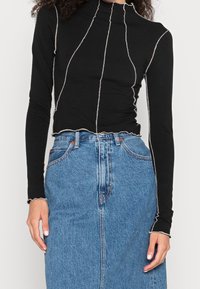 Svart långärmad topp med hög krage, med vit piping längs sömmarna och en scalloped nederkant, i kombination med en högmidjad denimkjol.