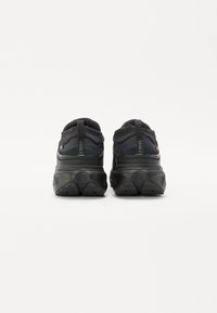 Chaussures de sport noires avec une tige en maille texturée, une semelle épaisse et rembourrée, et un marquage distinctif au niveau du talon. Comprend une languette pour un enfilage facile.