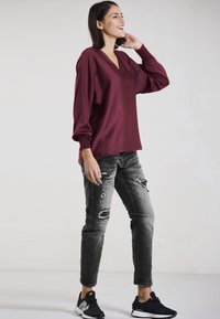 Burgundy V-neck blouse with balloon sleeves, paired with distressed gray jeans and black sneakers. Smooth fabric with a relaxed fit.