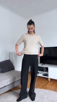 Bež pulover s kratkimi rokavi z teksturirano površino in okrasnim bisernim ovratnikom, kombiniran z ohlapnimi črnimi hlačami, stoji v sobi.