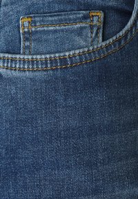 Tela de mezclilla de un azul oscuro que muestra un bolsillo trasero con costuras visibles. La textura es suave con ligeros patrones de desvanecimiento.