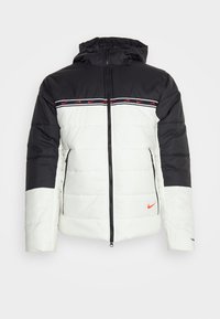 Vadderad jacka med svart överdel och vit nederdel. Har huva, dragkedja framtill och en röd Nike-logga på bröstet.