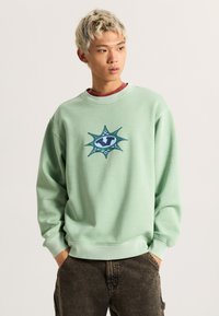 Felpa verde chiaro realizzata in tessuto morbido, con una grafica centrale blu e teal decorata con punti simili a stelle e accenti a scacchi.