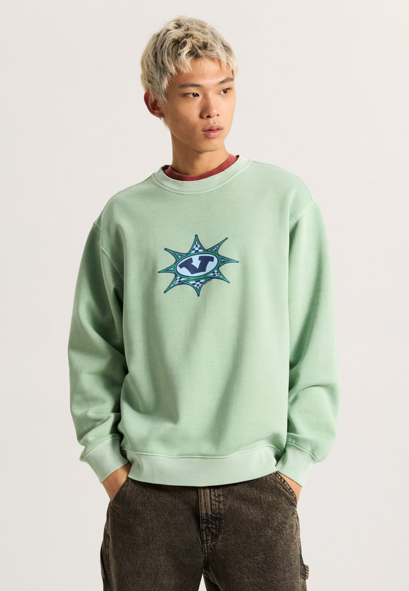 Felpa verde chiaro realizzata in tessuto morbido, con una grafica centrale blu e teal decorata con punti simili a stelle e accenti a scacchi.