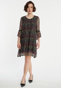 Robe noire transparente avec un motif zigzag multicolore ; possède des manches en cloche et un ourlet festonné. Portée avec des talons pointus noirs.