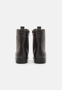 Un par de botines de cuero negro hasta el tobillo con cremalleras laterales y tiradores, mostrados desde la parte trasera sobre un fondo blanco.