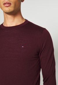 Bordeauxrotes Langarmshirt aus weichem Stoff mit Rundhalsausschnitt und einem kleinen, in Rot, Weiß und Blau gestickten Logo auf der Brust.