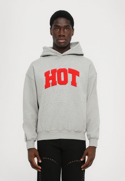Haut à capuche gris avec un grand texte rouge "HOT", capuche à cordon, poche kangourou et poignets côtelés. Porté avec un pantalon noir orné d'étoiles.