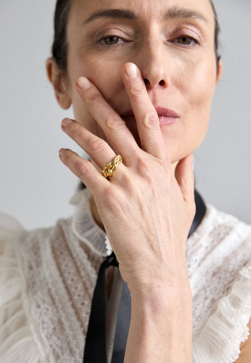Gold ring with an intricate design, worn on a hand resting on the chin. The person wears a ruffled white blouse with a black ribbon.