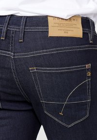 Jeans de denim azul oscuro con costuras amarillas en contraste, que presentan un diseño de bolsillo trasero, etiqueta marcada "KOROSHI" y acentos de hardware distintivos.
