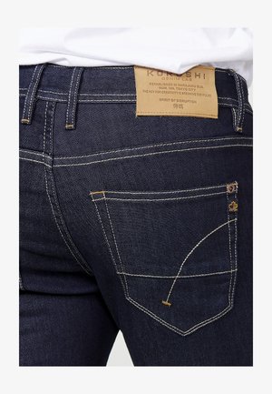 Jeans de denim azul oscuro con costuras amarillas en contraste, que presentan un diseño de bolsillo trasero, etiqueta marcada "KOROSHI" y acentos de hardware distintivos.