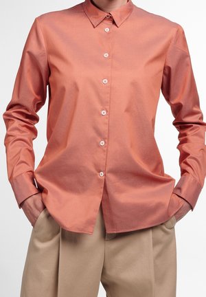 Person trägt ein lachsfarbenes, langärmeliges Hemd mit Knöpfen und beige Hose, Hände in den Taschen, vor einem schlichten weißen Hintergrund.