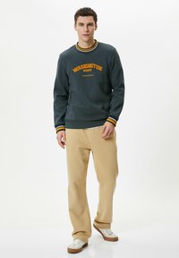 Mørkegrøn sweatshirt med gule og sorte striber ved crew-neck, med "Washington State" broderet i orange. Beige afslappede bukser og sneakers.