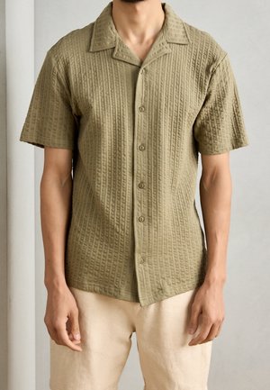 Mann trägt ein strukturiertes olivgrünes Kurzarm-Button-up-Hemd und beige Shorts, steht vor einem hellgrauen Hintergrund.