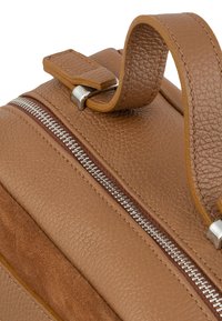 La mochila de cuero marrón y ante presenta un acabado texturizado, un zipper plateado y un asa cosida para un fácil transporte.