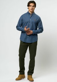 Man in een blauw overhemd met knopen, donkergroene broek en tan-kleurige laarzen, staand tegen een effen lichte achtergrond.