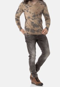 Maglietta a maniche lunghe e scollo a V beige con macchie scure, abbinata a jeans slim-fit grigi e stivali da escursionismo marroni.