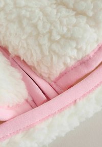 Lähikuva valkoisesta pörröisestä kankaasta, jossa on pehmeän vaaleanpunaiset reunat ja hihnat, esittäen tekstuuria ja ompelun yksityiskohtia.