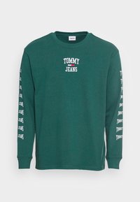 Vihreä pitkähihainen t-paita, jossa on valkoinen "TOMMY JEANS" -logo rinnassa ja toistuva teksti molemmissa hihoissa. Valmistettu pehmeästä puuvillamateriaalista.