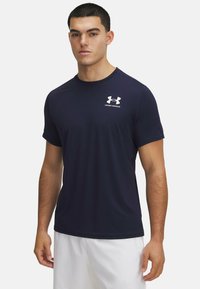 Tamsiai mėlynas trumpomis rankovėmis sportinis marškinėlis, pagamintas iš lengvos medžiagos. Kairėje krūtinės pusėje yra balta „Under Armour“ logotipo etiketė. Lygus paviršius.