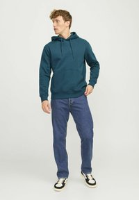 Türkise Kapuzenjacke mit Kängurutasche und verstellbarem Kordelzug, kombiniert mit blauen Jeans und schwarz-weißen Turnschuhen.