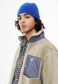 Mladostnik s kodrasto frizuro, oblečen v modro pleteno kapo, bež flis jakno z modrim žepom in črno-belo karirasto srajco, obrnjen nekoliko levo.