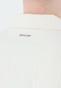 Camisa blanca texturizada con diseño acanalado y cuello doblado. Logo PXP en dorado metálico bordado en la parte trasera cerca del escote.
