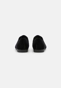 Crne baršunaste cipele na slip-on s niskom petom, zaobljenim stražnjim dijelom i teksturiranom tkaninom. Imaju glatku podlogu s minimalnim akcentima.