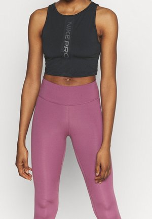 Kobieta w czarnym krótkim topie Nike Pro i wysokich różowofioletowych legginsach, stojąca na jednolitym jasnym tle.
