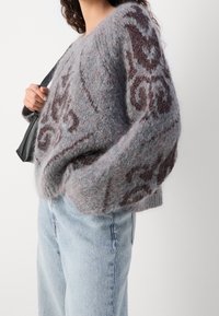 Žena v chlupatém šedém svetru s tmavě fialovými vzory, světle modrých džínách a držící černou kabelku přes rameno na bílém pozadí.