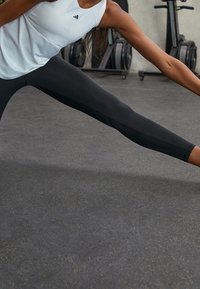 Biała kamizelka bez rękawów z czarnym logo, w połączeniu z czarnymi dopasowanymi legginsami. Gładki materiał i sportowy krój, rozciągający się na ciemnej podłodze w siłowni.
